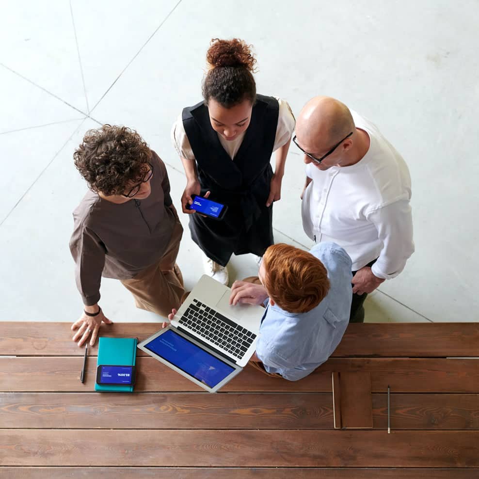 Four people are standing around a wooden table having a discussion. One person is holding a smartphone, another is using a laptop. They appear to be collaborating on a project. The table has a few items on it, such as a notebook and a pen.