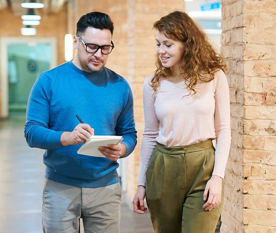 A man in a blue sweater and gray pants is writing on a notepad while walking alongside a woman in a light pink long-sleeve shirt and olive green pants. They are in a hallway with exposed brick walls, engaging in conversation.