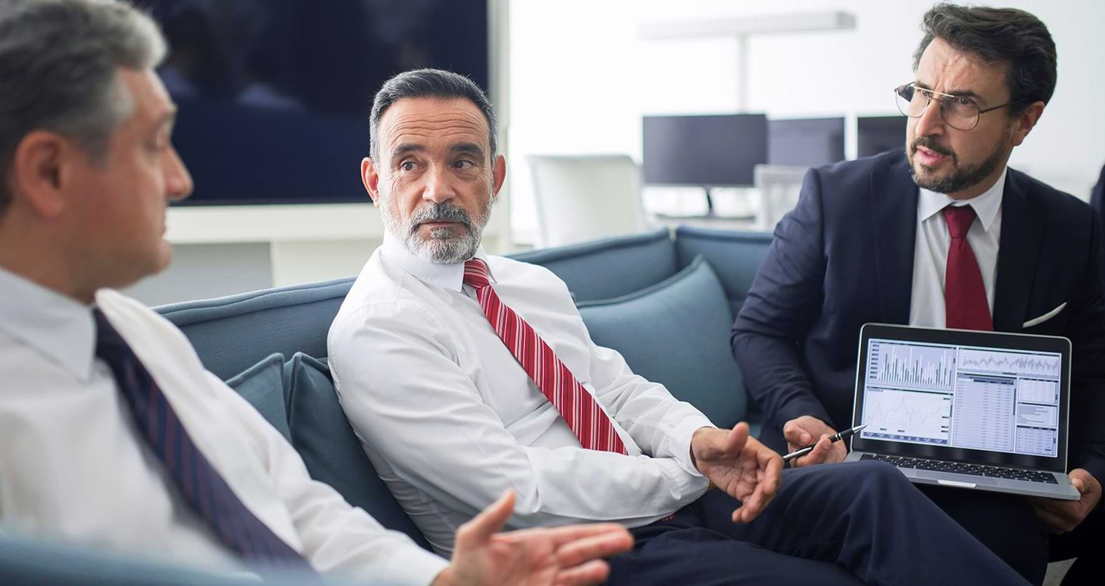 Three men in business attire are having a serious discussion in a modern office. One is holding a laptop showing graphs and data. They appear to be analyzing information or brainstorming ideas. Modern workstations and a large screen are visible in the background.