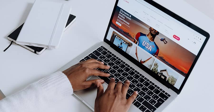 A person wearing a white long-sleeved shirt is typing on a silver laptop displaying a colorful website. The laptop is on a white desk with an open notebook and a pen beside it.