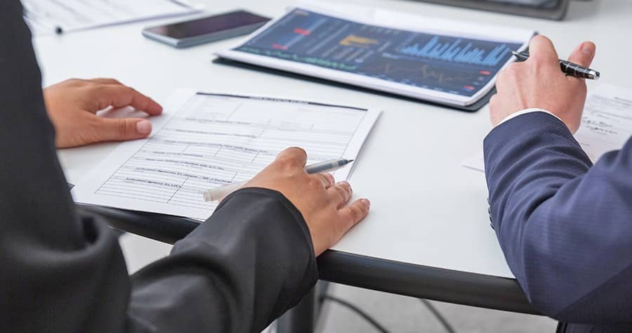 Two individuals in business attire are seated at a desk. One person is holding a pen and a document, while the other points to a chart on a tablet displaying graphs and data. A phone and another tablet are also on the desk.